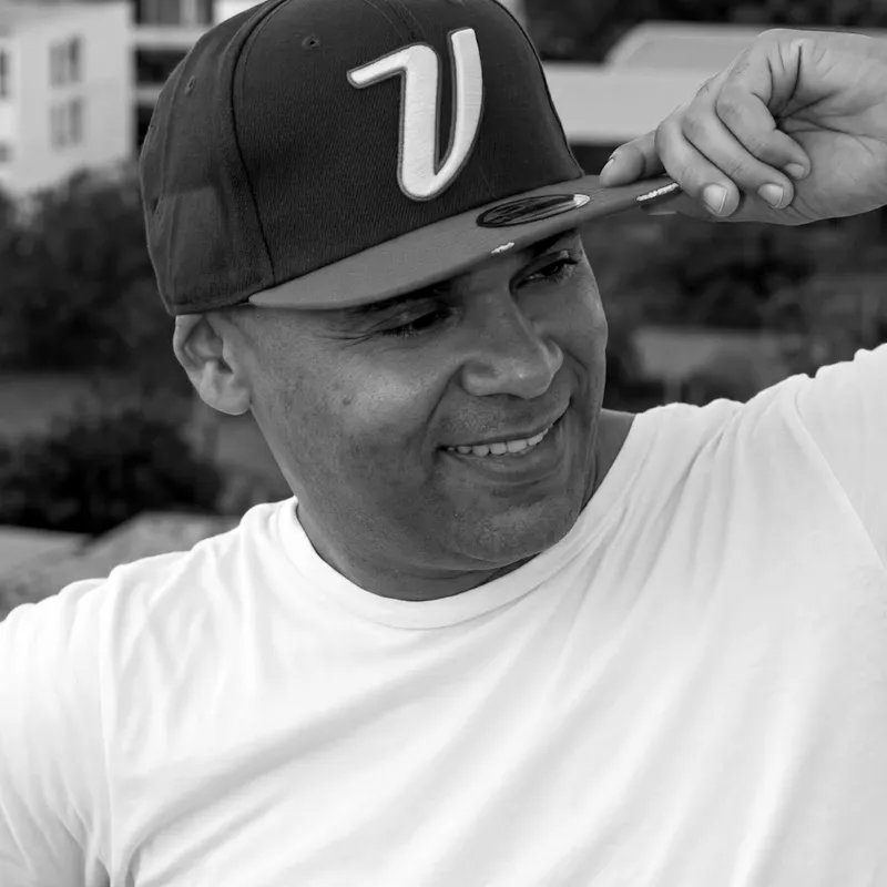 Retrato en blanco y negro de Robert Virona con gorra, sonriendo, dentro de un anillo naranja